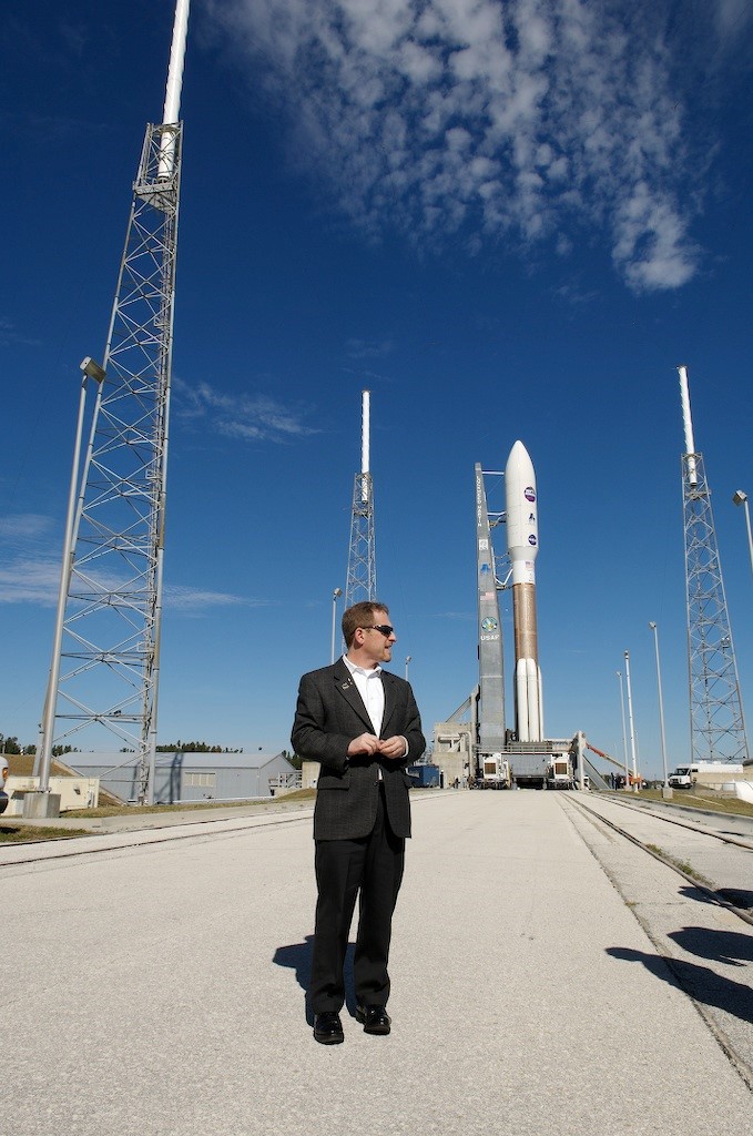 S. Alan Stern standing outside, with a rocket in the distance behind him. Photo courtesy of NASA.