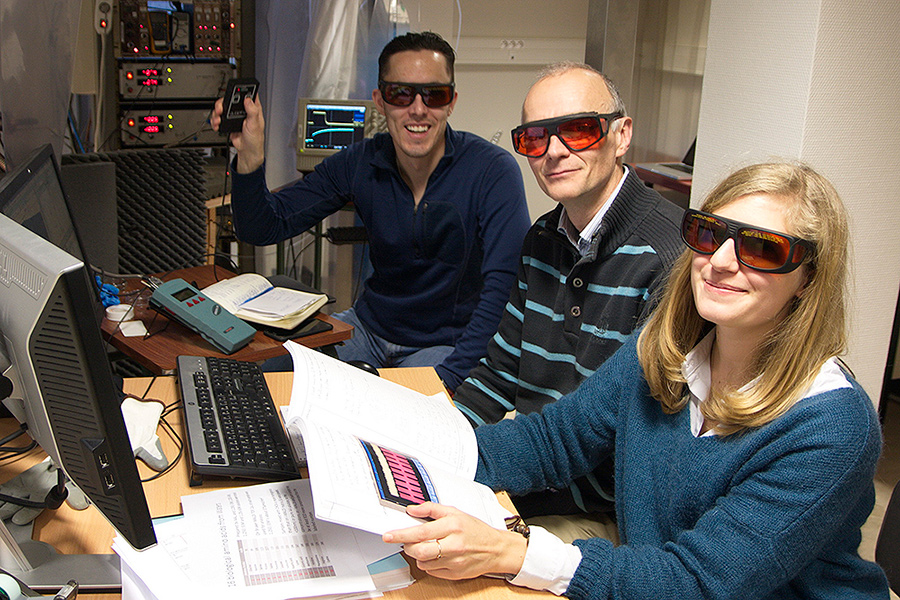 Ricardo Arevalo and two co-researchers - a man and a woman - sitting at a computer, A rack of equipment is in the background and all three are wearing red-orange tinted protective glasses.