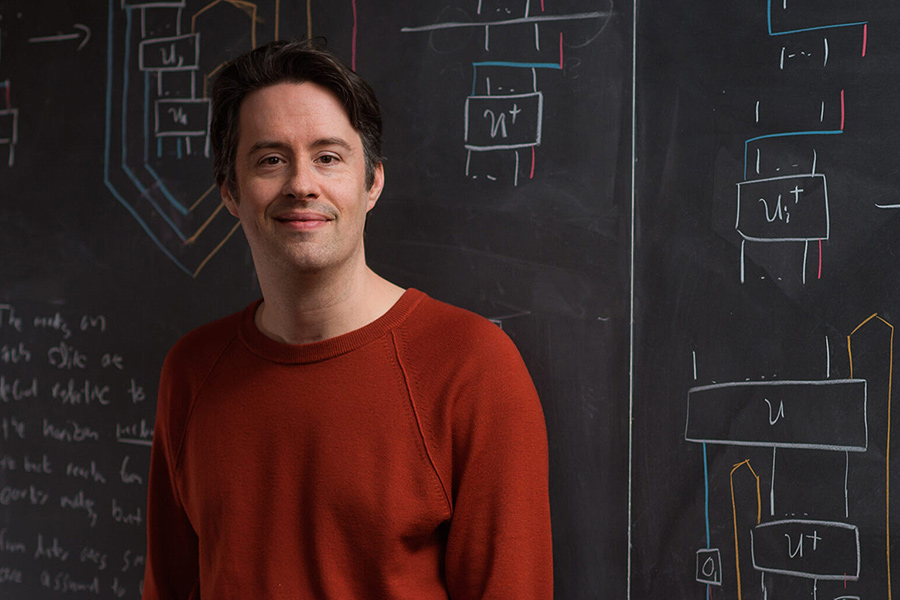 Daniel Harlow standing in front of a blackboard with colorful diagrams on it. Credit: MIT