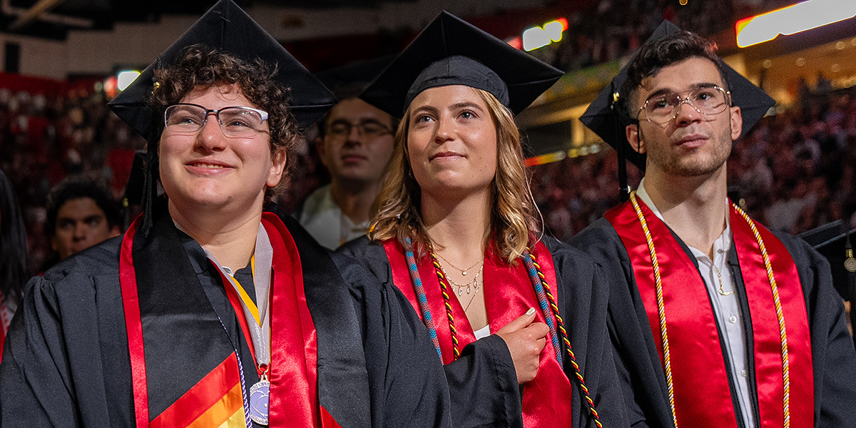 A row of three undergraduate students, two women and a man, at graduation, wearing regalia. Credit: Mark Sherwood.