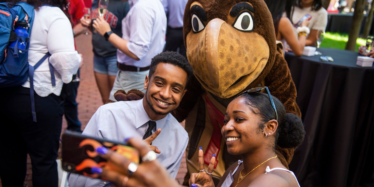 A young Black man and young Black woman at an outdoor reception, taking a selfie with the Testudo mascot. Photo by Lisa Helfert.