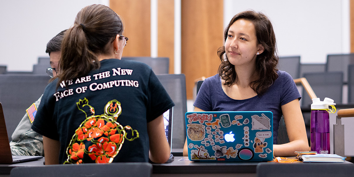 Two young women in a lecture hall working at their laptops. One is wearing a shirt that says, We are the new face of computing.