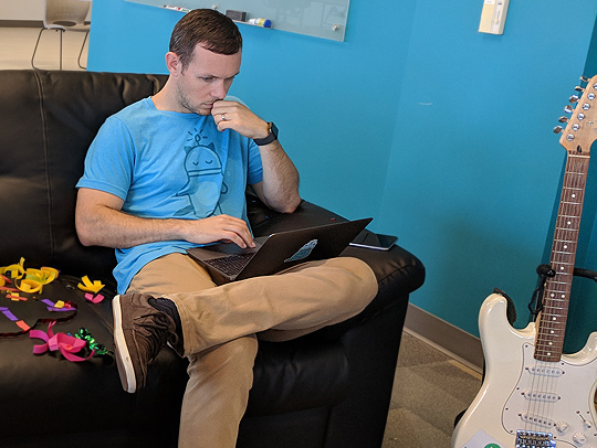 Paul Capriolo sitting in a bright blue room on a black leather couch. He is working on his laptop and a white electric guitar is on a stand next to him. Photo courtesy of Paul Capriolo.