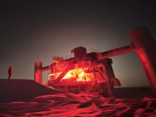 A night shot of Icecube Base in Antartica. The light from within the blocky, industrial-style structure is yellow and orange. A person is standing outside nearby. Credit: IceCube Collaboration-NSF-Lily Le-Shawn Johnson-ESO-S Brunier.