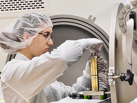Sarah Waldych, wearing a special gown, hairnet and gloves, in the final stages of preparing for a new sample to be tested in a very sensitive thermal experiment inside the vacuum chamber.