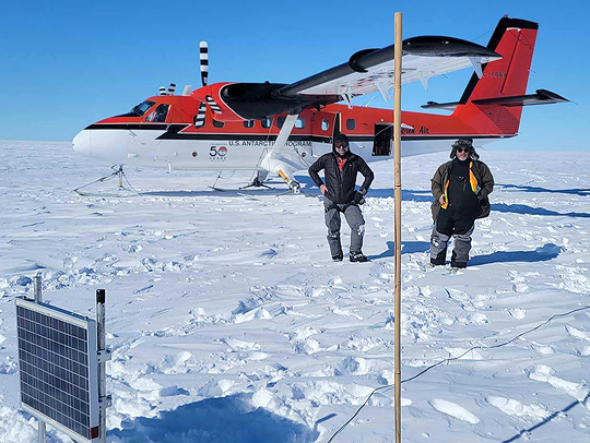 Associate Professor Nick Schmerr, left, with pilot Troy McKerral, behind a GPS station with a solar panel, recording unit, and antenna. The bamboo pole helps the retrieval team find the equipment, since the ice shelf can move a meter per day.