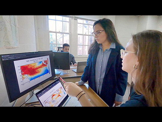 Letterboxed video still of Professor Maria Molina and a female student checking weather forcast simulations on a computer screen. A male student is working at his laptop in the background.