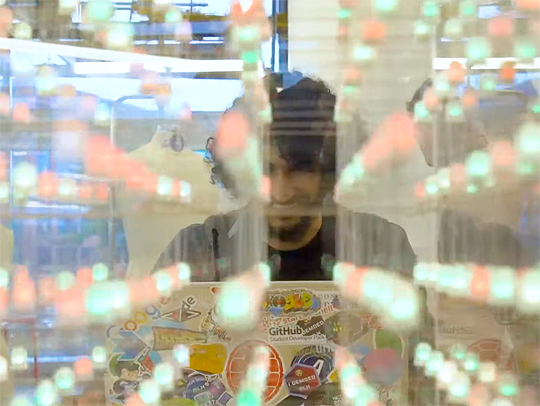 A photo of a male student behind his laptop. There are many tech-oriented stickers on his laptop. We are viewing him through a grid of green and red lights in soft focus.