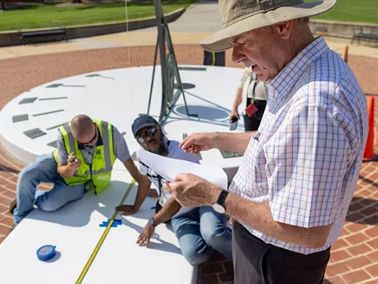 Andy Harris consults plans as men in hi-viz vests repair the sundial behind him. Credit: WJLA 7