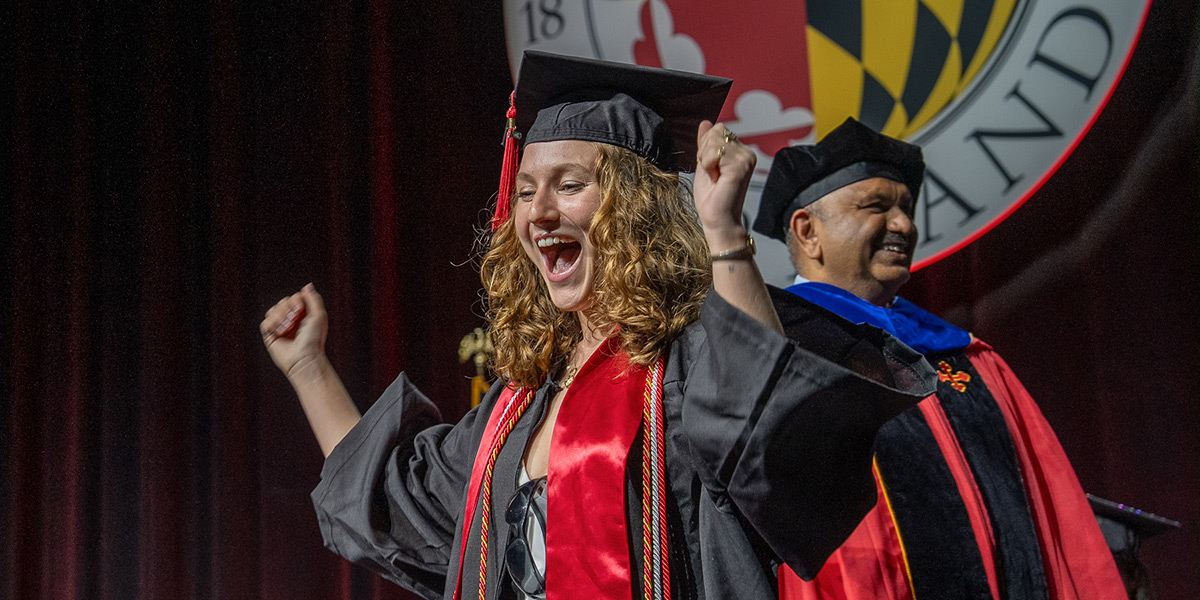 A young woman in regalia stops to cheer as she crosses the stage at the undergraduate commencement. Dean Amitabh Varshney, also in regalia, stands smiling in the background, and above there heads is a large, lit-up sign of the University of Maryland seal Credit: Mark Sherwood.