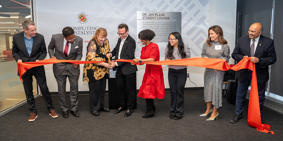 At the dedication of the Jan Plane Lounge, a group of people holds a large red ribbon as Jan cuts it. Behind them is a wall decorated with the Computing Catalyst logo and a plaque with the name of the lounge and information about Jan. Credit: Mark Sherwood.