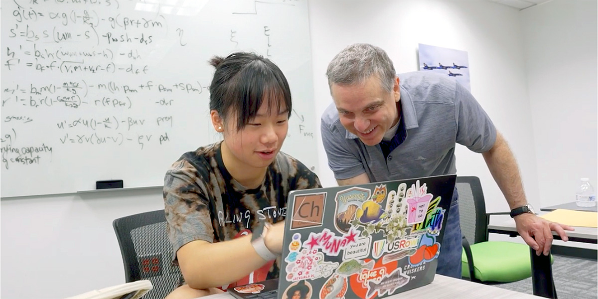 UMD mathematics Lecturer Todd Rowland leaning in to look a female high school student's laptop screen. Her laptop lid is covered with colorful stickers. Behind the pair is a glassboard full of equations. Credit: Mark Sherwood.