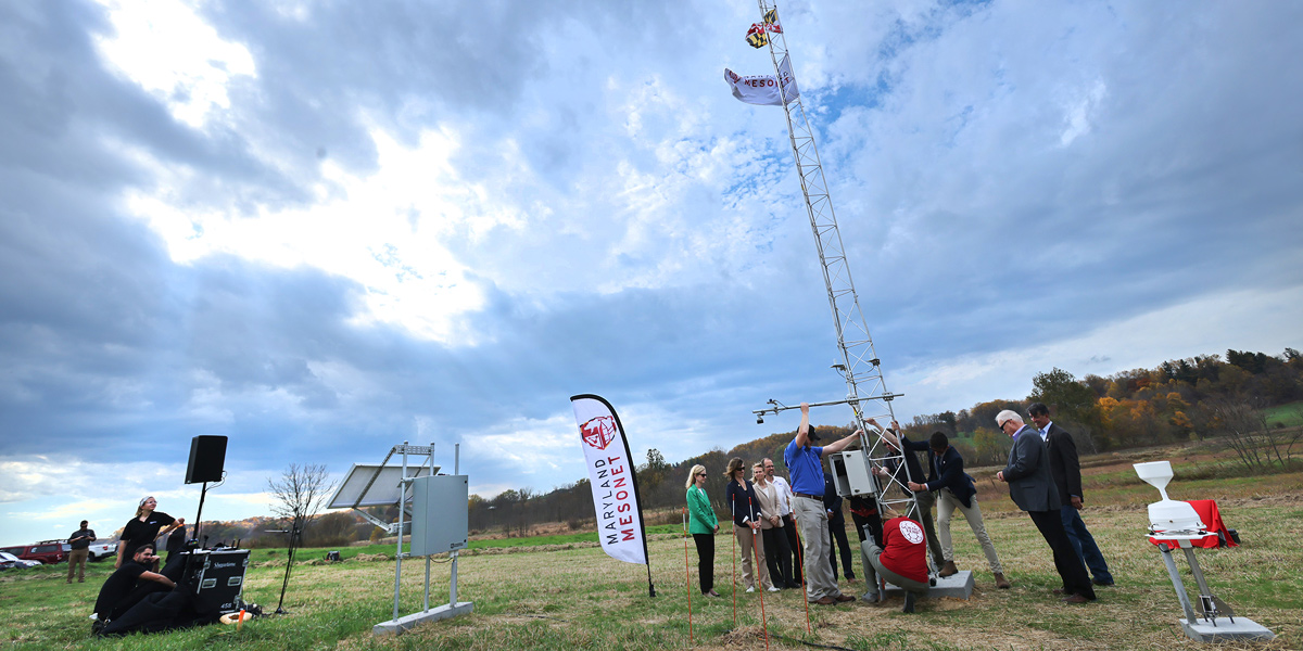The Maryland Mesonet team sets up the first tower of the state's atmospheric monitoring system. The slim metllic tower, mounted in a grassy field, looks somewhat like a radio tower and has Maryland and US flags on it.