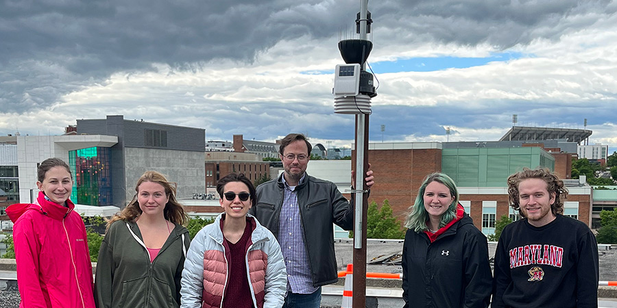 A group photo of the Mesoterps with their newest weather station on the roof of the A.V. Williams Building. The sky is filled with dramatic clouds. Campus buildings are visible in the distance.