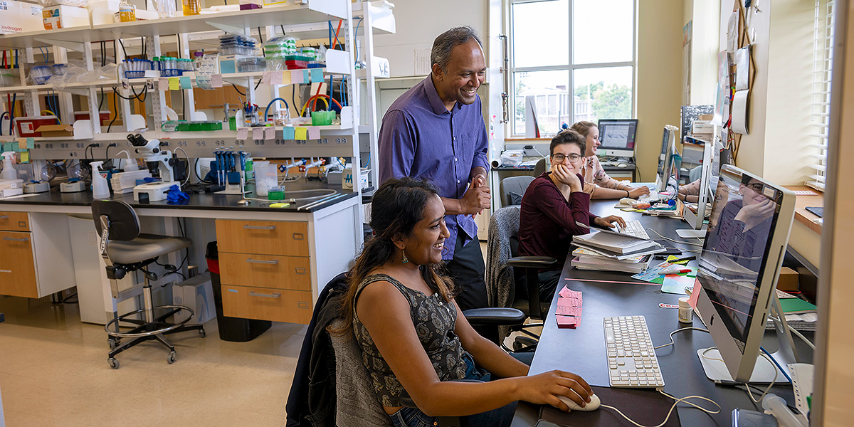 CBMG professor Antony Jose in his lab with three female students. The three women are sitting at a long counter, each at an iMac computer. Professor Jose is standing between two of them, looking at infomation on a screen. Everyone is smiling. Lab workbench stations and stocked shelves are in the background. Credit: Lisa Helfert.