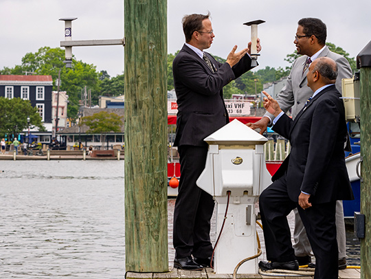 Professor Tim Canty demonstrating a Hydronet sensor for CMNS ean Amitabh Varshney and UMD President Darryll Pines on a dock in Annapolis. The Chesapeake Bay and historic rowhomes are in the background. Credit: Mark Sherwood.