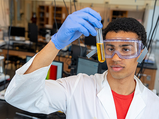 Jamil Yacoubou Adam in a lab, wearing a white lab coat, blue gloves, and eye protection. He is holding a test tube with an orange liquid in it. Credit: Mark Sherwood