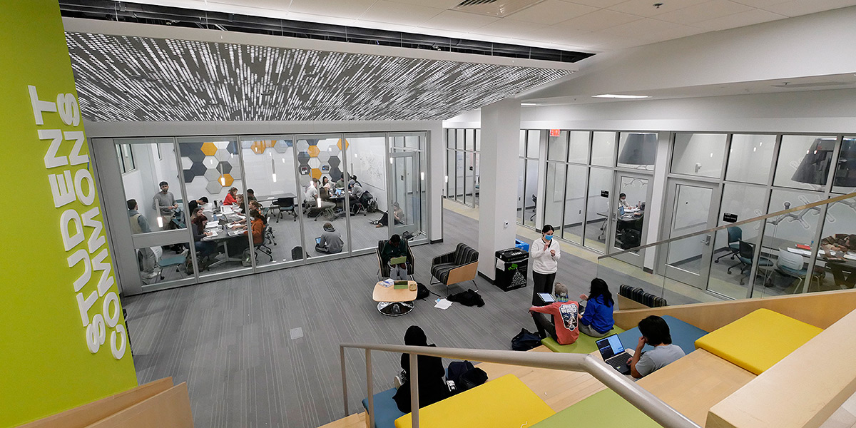 A wide angle photo looking down from the raised entrance of the new Chemistry Student Commons. Students are gathered on open step seating and at tables, and larger groups are in huddle rooms along the walls. The design is modern with a silver, pale wood, gold, and lime green color palette.