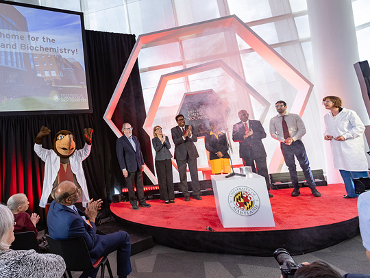 Testudo, wearing a lab coat, cheers on VIPs and spekers standing on a red-carpeted stage. A lit double hexagon sculture forms the backdrop. Credit: John T. Consoli.