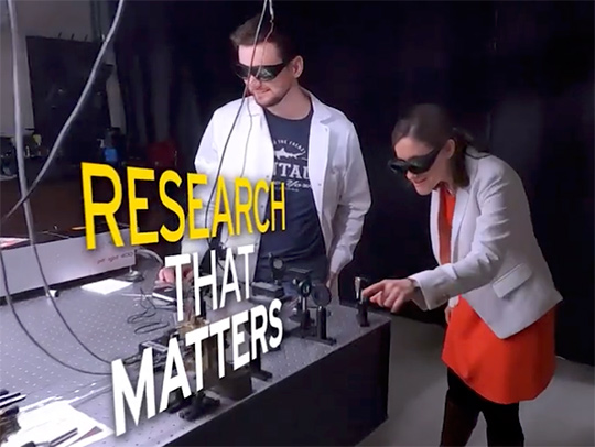 A still from the video with the floating title 'Research That Matters.' A grad student and professors are at an anti-vibration table on which equipment is mounted. Both are wearing lab coats and dark eye protection.