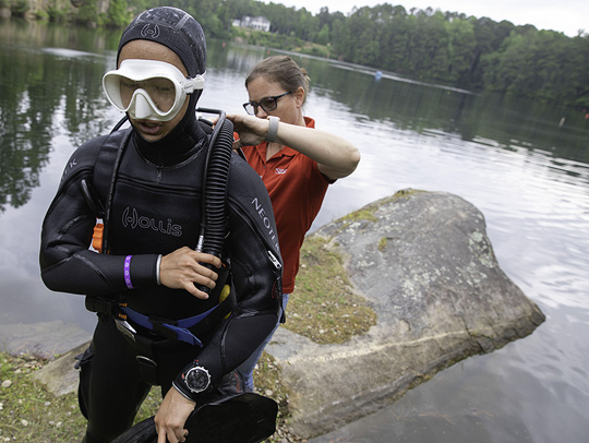 Grant Dong, dressed in full SCUBA gear, is helped by another researcher while standing on the shore of a lake or bay.