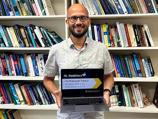 Pratyush Tiwary standing in front of a bookcase, holding his laptop.