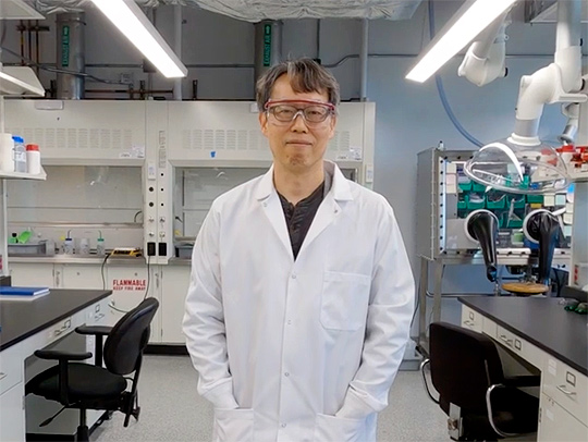 Sang Bok Lee wearing a lab coat, hands in its pockets, stnding between two bench stations in his lab.