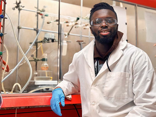 Solomon Attionu, wearing a white lab coat, blue gloves, and eye protection sitting in front of a rack of tubes and flasks.