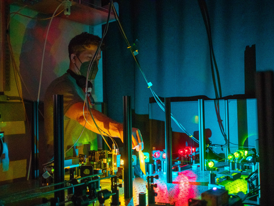 Colenso Speer in a darkened room lit by colored lasers. Speer is highlighting a beam path on his group's custom-built instrument for super-resolution single-molecule localization microscopy. This microscope enables his team to visualize proteins within large tissue volumes with nanoscale image resolution.