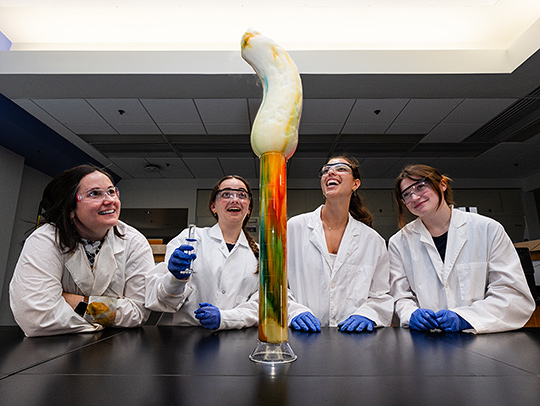 A group of 4 young women in lab coats looking up at colored foam erupting from a large flask. Credit: Mark Sherwood