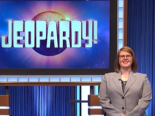 Ciara Donegan on the Jeopardy! set in front of a large screen displaying the show's logo. Photo courtesy of Ciara Donegan.