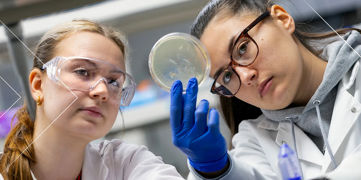 Two young women in lab coats at a lab bench looking at a petri dish. Credit: John T. Consoli.