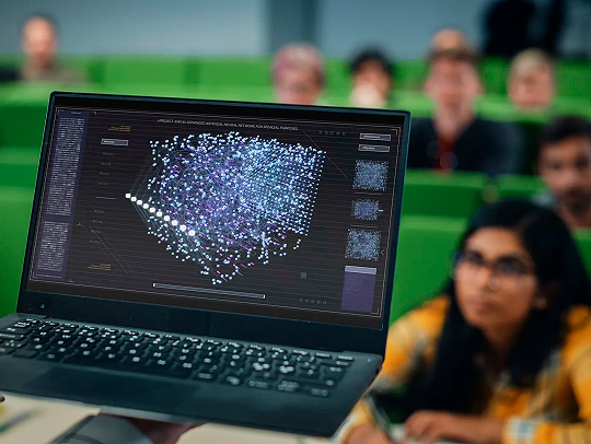 A small laptop resting on someone's palm with data on the screen. There are students at desks in a lecture hall in soft focus in the near distance. Credit: AdobeStock