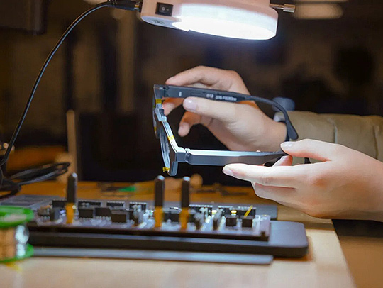 Hands holding a pair of glasses that look something like thick Ray-Bans. Electronig equipment is on the table beneath them.