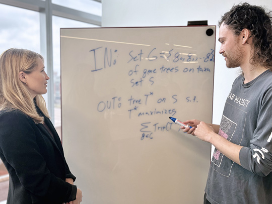 A young man and a young woman stading in front of a whiteboard with equations on it.