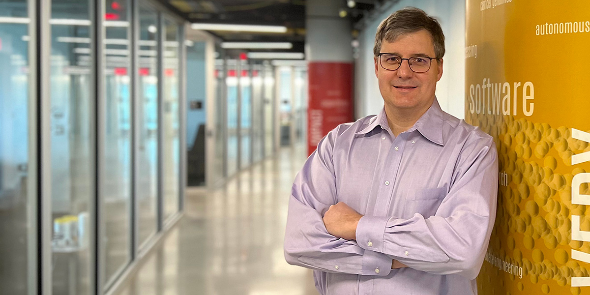 Computer Science Professor and Chair Mihai Pop leaning against a decorated pillar in the Iribe Center. A long, slightly curved hallway of glass-walled rooms is in soft focus in the background. Credit: Tom Ventsias