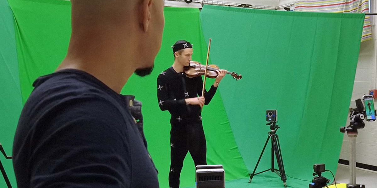 Snehesh Shrestha, foreground, watches a musician. The musician is wearing a motion capture suit and playing the violin while being recorded by multiple cameras.
