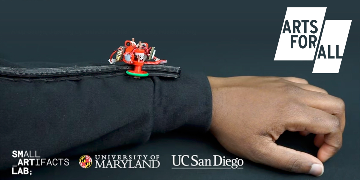 A closeup shot of a forearm resting on a table. There is a narrow track on the sleeve. Making its way down the track is a little red robot. Logos for Arts for All, the Small Artifacts Lab, the University of Maryland, and UC San Diego are in the margins of the photo.