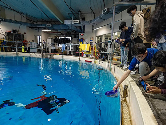 A group of undergraduate students testing a bio-inspired robot that could address an environmental challenge. Some are at the edge of a pool and one of them is a diver in the water.