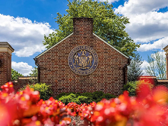 UMD's Founders' Gate, a brick structure with a metal plaque on it bearing the Maryland seal. Flowers are in the foreground.