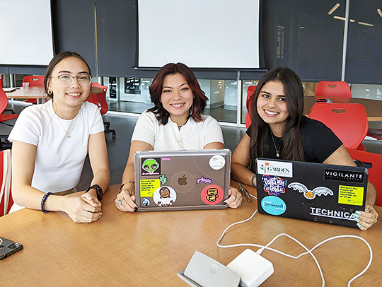 A trio of young women sitting at a classroom table. Two of them have laptops in front of them, both of which have multiple stickers on their cases.