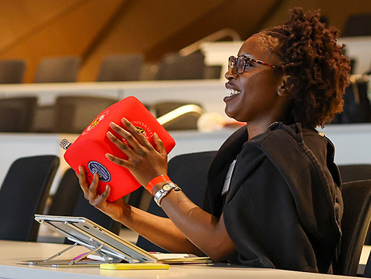 A smiling young woman holding a prototpe object that looks like a soft red box. A tablet computer is on the desk in front of her.