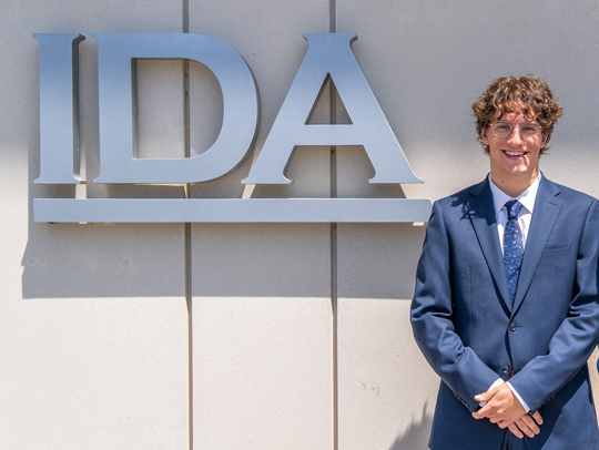 CS student Hayden Earlwine, wearing a suit, poses in front of the  Institute for Defense Analyses logo