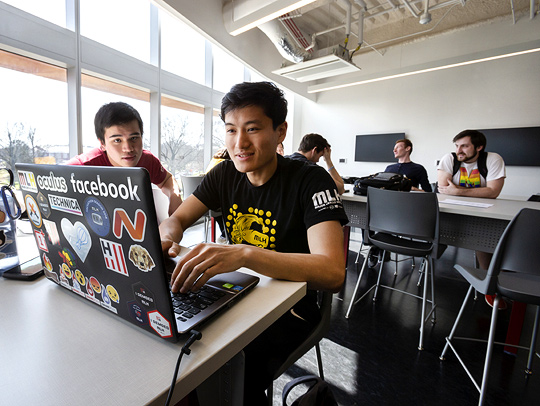 UMD CS students in a classroom in the Iribe Center. In the foreground, two oung men are working on a laptop whose cover is covered with stickers. Credit: John T. Consoli.