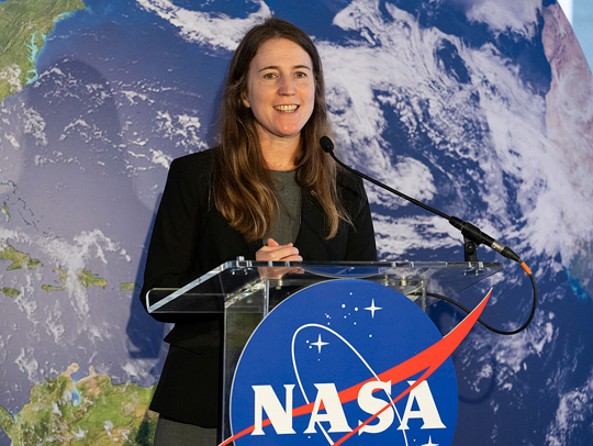 Katherine Calvin speaking at a lucite podium with NASA logo on it. Behind her is a giant globe.