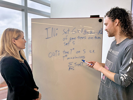 Erin Molloy standing on the left side of a whiteboard containing data, talking to a graduate student on the right.
