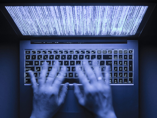 Stock art of motion-blurred hands over a laptop keyboard. The laptop screen has computer code on it. The lighting is blueish-purple.