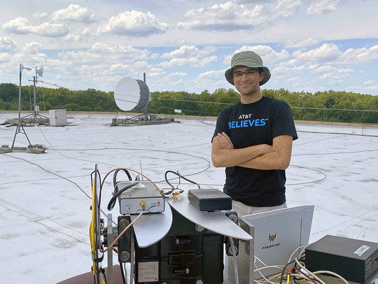 Neil Sorkin on a roof with computer equipment. A blue sky and fluffy clounds are in the background.