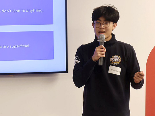 A male student presenting at the Pitch Perfect event. He's holding a microphone and standing in front of a screen.
