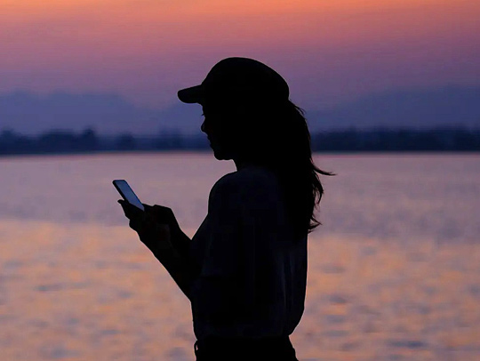 Stock art of a woman in silhouette against a vibrant sunset over water, checking her phone.